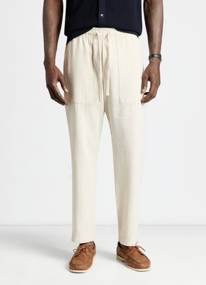 Uomo che indossa pantaloni beige con coulisse, scarpe marroni stringate e una camicia scura a maniche corte, in piedi su uno sfondo bianco uniforme.
