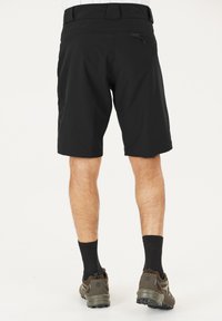 Man in zwarte shorts met een achterzak met rits, zwarte sokken en olijfgroene sportschoenen, staand op een witte achtergrond.