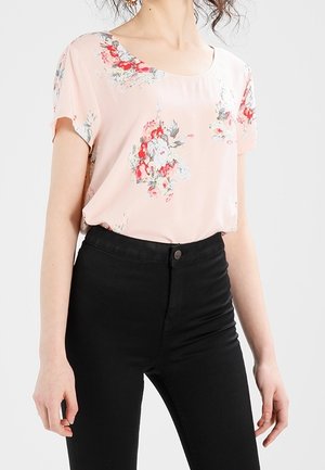 Femme portant un chemisier à manches courtes fleuri rose clair, rentré dans un pantalon noir taille haute, debout sur un fond blanc.