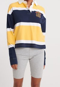 Maglietta da rugby a maniche lunghe nei colori blu navy, giallo e bianco a righe, con colletto e logo ricamato; abbinata a pantaloni corti da ciclismo grigi.