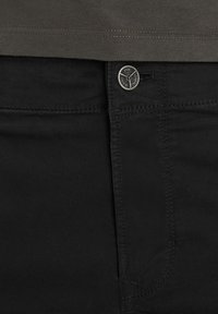 Gros plan sur un jean en denim noir avec des coutures visibles, une poche avant et un bouton métallique marqué sous une chemise vert olive foncé.