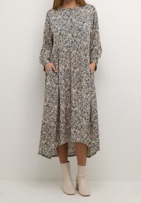 Abito floreale con maniche lunghe, colletto alto e tasche. Presenta motivi viola, verdi e gialli su uno sfondo chiaro. Di lunghezza media e fluente.