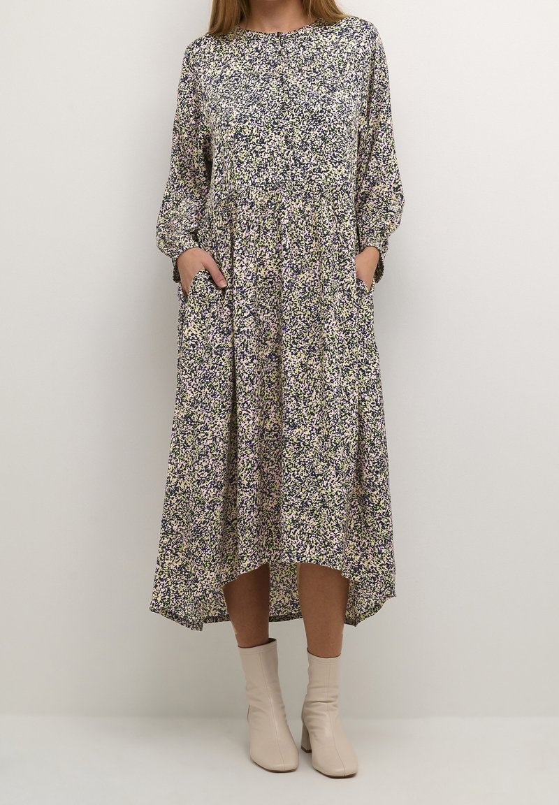 Abito floreale con maniche lunghe, colletto alto e tasche. Presenta motivi viola, verdi e gialli su uno sfondo chiaro. Di lunghezza media e fluente.
