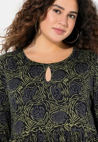 Černý top s zeleným abstraktním vírem a květinovým vzorem. Má výstřih ve tvaru klíčové dírky a dlouhé rukávy, vyrobený z texturované tkaniny.