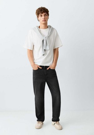 Junger Mann steht mit den Händen in den Taschen, trägt ein weißes T-Shirt, schwarze Jeans, beige Sneakers und einen grauen Pullover über den Schultern.