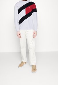 Hellegraues Langarmhemd mit diagonalen schwarzen, roten und weißen Streifen. Kombiniert mit weißen Hosen und tanfarbenen Sneakern.