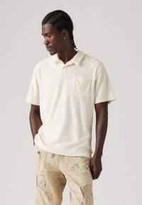 Polo tričko s krátkym rukávom v off-white farbe, vyrobené z mäkkého materiálu, s klasickým golierom a náprsné vrecko na ľavej strane. Nosené s vzorovanými béžovými cargo nohavicami.