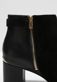 Bottine en cuir noir avec talon bloc, fermeture éclair dorée sur le côté ornée du logo "ck" et partie supérieure en daim.