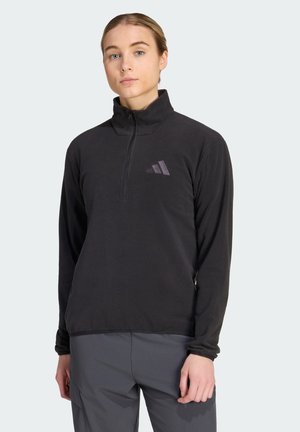 Jong persoon met licht haar die een zwarte quarter-zip fleece met een subtiel logo en donkergrijze broek draagt, kijkt recht vooruit met een neutrale uitdrukking.