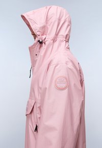 Άτομο που φοράει ροζ αδιάβροχο με κουκούλα, φερμουάρ στον ψηλό γιακά και στρογγυλή κεντημένη ετικέτα στο πάνω μέρος του χεριού, κοιτάζοντας προς τα αριστερά.