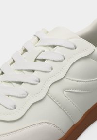 Κοντινό πλάνο λευκού αθλητικού παπουτσιού με λευκά κορδόνια, υφασμένη ραφή και καφέ σόλα, που δείχνει λεπτομερώς τον σχεδιασμό του παπουτσιού.