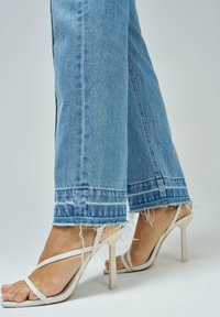 Jeans de tiro ancho en azul claro con bordes deshilachados, combinados con sandalias de tacón alto en color crema con tiras. Enfoque en la textura de la tela y los detalles del dobladillo.