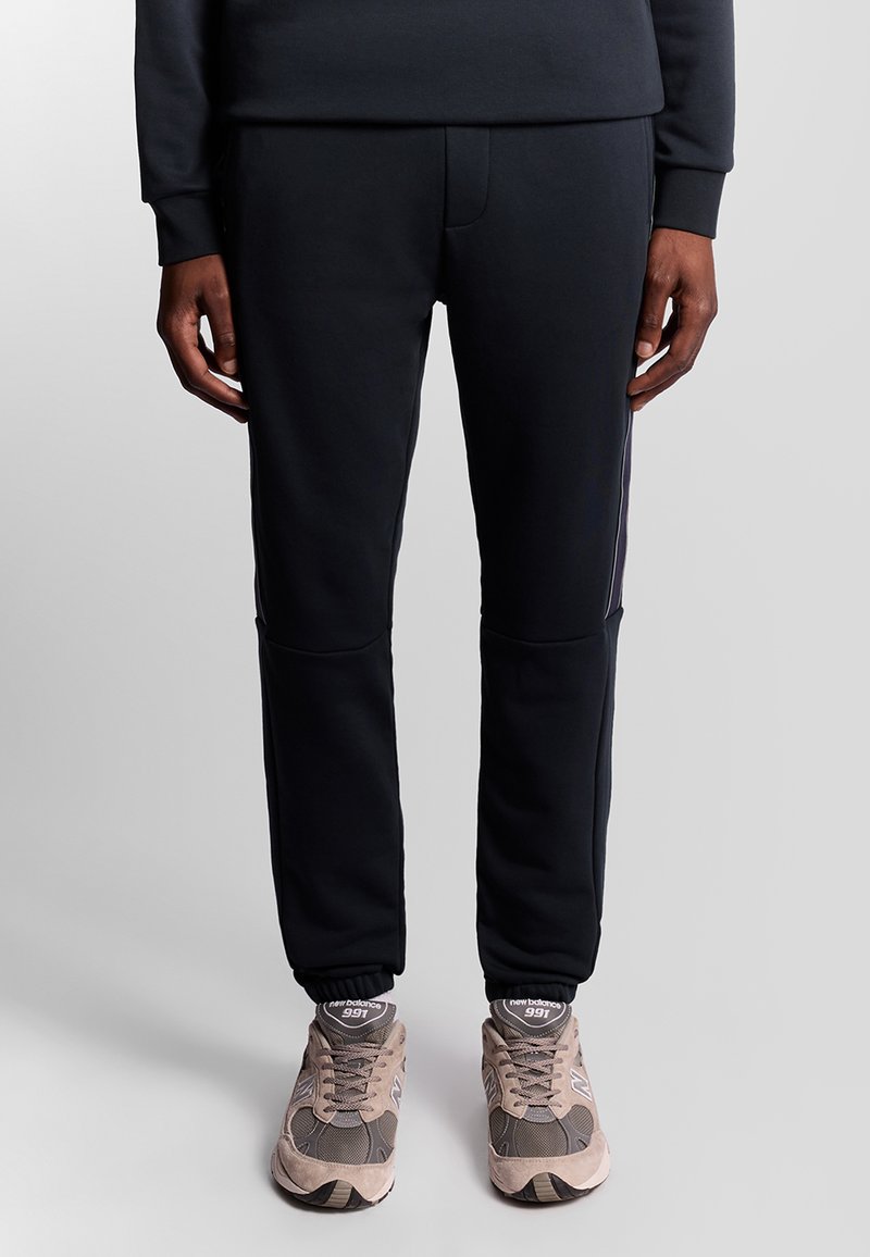 Pantaloni jogger navy in tessuto morbido con polsini a coste, dotati di strisce laterali e una tasca frontale. Abbinati a sneakers beige.