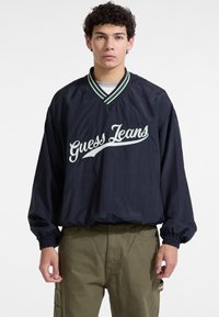 Jūrinis vėjo nepralaidus paltas su žaliais dryžiais ant apykaklės, su baltu siuvinėtu "Guess Jeans" tekstu per krūtinę ir elastinėmis rankovėmis.