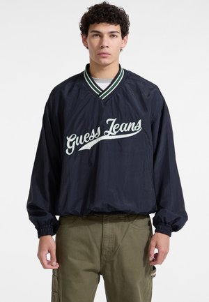 Chaqueta cortavientos de color navy con un cuello a rayas verdes, que presenta el texto bordado en blanco "Guess Jeans" en el pecho y puños elásticos.