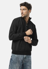 Zwarte zip-neck trui met ribbeltextuur en lange mouwen, met een aansluitende taille en manchetten, gecombineerd met lichte denim jeans.