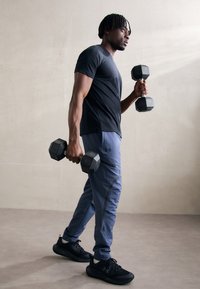 T-shirt de sport noir et survêtement bleu clair, tenant des haltères noirs. Comprend une poche zippée sur le pantalon et des chaussures de sport noires.