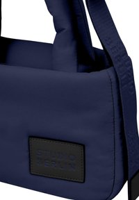 Borsa imbottita blu navy con un'etichetta nera testurizzata con la scritta "STUDIO BERLIN" in rilievo. Include una tracolla intrecciata con dettagli a motivo.