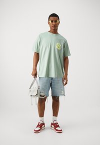 Camiseta de algodón verde claro con un gráfico circular amarillo, combinada con pantalones cortos de mezclilla desgastados y zapatillas rojas y blancas; bolso cruzado gris.