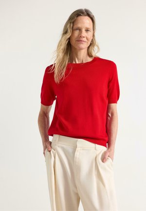 Donna con lunghi capelli biondi che indossa un maglione rosso a maniche corte e pantaloni crema a gamba larga, in piedi con le mani nelle tasche.