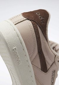 Gros plan sur le talon d'une chaussure Reebok beige et marron, montrant la semelle texturée et le panneau arrière en daim avec les logos de la marque sur la semelle et la languette du talon.