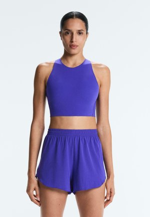 Sportliche Frau mit dunklen Haaren in einem lila Crop-Top mit hohem Kragen und passenden Laufshorts mit elastischem Bund, stehend vor einem schlichten hellen Hintergrund.