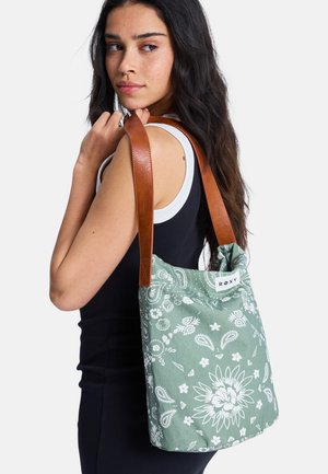 Kobieta z długimi ciemnymi włosami, ubrana w czarny bezrękawnik, niesie zielono-białą torbę typu tote z kwiatowym wzorem i brązowymi skórzanymi paskami.