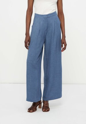 Γυναίκα που φοράει μπλε παντελόνι με ψηλή μέση και φαρδιά μπατζάκια και καφέ σανδάλια με τακούνι, στέκεται μπροστά σε λευκό φόντο.