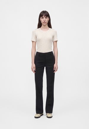 Femme debout, visage vers l'avant, portant un haut beige à manches courtes, un pantalon noir droit et des chaussures beiges sur un fond blanc uni.