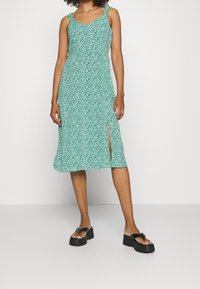 Vestido floral verde con tirantes finos, escote en V y abertura lateral. Hecho de una tela ligera con un corte relajado. Sandalias de plataforma negras.