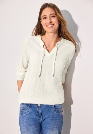 Blouse texturée vert clair avec un décolleté en V et des cordons, associée à un jean bleu. La blouse présente des détails froncés sur les épaules.