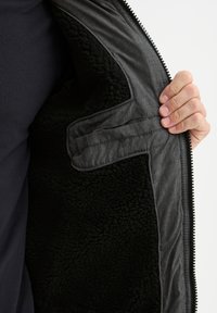 Sort fleece-foring jakke med et gråt yderstof. Indersiden har en glat tekstur og en lomme detalje. Lynlås lukning.