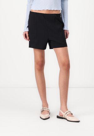 Black shorts with a tailored design, featuring front pleats and a smooth texture, paired with pale pink pointed-toe slingback shoes.