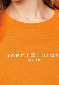 Camisa de manga corta naranja con un tejido texturizado y un logo blanco que dice "TOMMY HILFIGER EST. 1985" en el área del pecho.
