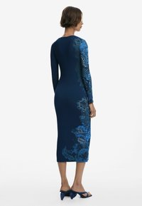 Langärmliges, marineblaues Kleid mit blauen Blumenmuster auf einer Seite, figurbetontes Design, knielang, gerippte Textur, kombiniert mit blauen Absatzsandalen.