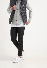 Chaleco acolchado negro sobre una camisa de botones gris, combinado con jeans negros de corte ajustado y zapatillas blancas con tiras de velcro.