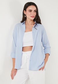 Light blue, textured shirt with buttons and rolled sleeves, worn open over a white crop top. Paired with white drawstring pants.