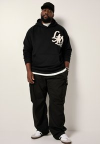 Schwarzer Kapuzensweater mit weißem Logo, kombiniert mit schwarzen Cargo-Hosen und weißen Sneakers. Das Model trägt eine schwarze Mütze und hat einen Bart.