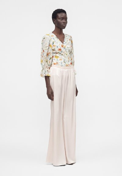 Γυναίκα που φοράει μπλούζα με μακριά μανίκια και floral σχέδιο και ψηλόμεσο, φαρδύ, κρεμ παντελόνι, στέκεται μπροστά σε απλό λευκό φόντο.