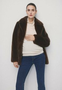 Vrouw in een donkerbruine nepbontjas, beige coltrui en blauwe spijkerbroek, staand tegen een eenvoudige lichte muur, met één hand op haar buik.