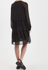 Černé šaty s puntíky, dlouhými rukávy a vrstveným designem. Vyrobeno z průsvitné tkaniny s jemnou strukturou a středně dlouhým střihem. Černá obuv.