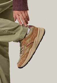 Sneaker in suede color tan e verde oliva con suola spessa, accenti viola a contrasto e design con lacci, posizionata su uno sfondo chiaro.
