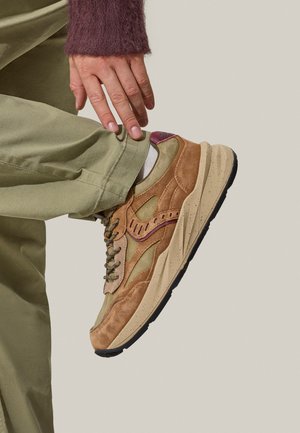 Sneaker in suede color tan e verde oliva con suola spessa, accenti viola a contrasto e design con lacci, posizionata su uno sfondo chiaro.