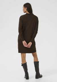 Robe tricotée marron à manches longues, col montant, et coupe décontractée, associée à des bottes en cuir noires jusqu'aux genoux et à une texture lisse.