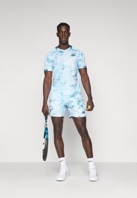 Sinine tie-dye spordivorm koosneb lühikeste varrukatega V-kaelusega ülemisest osast ja sobivatest lühikestest pükstest. Peal on valged sneakers ja käes tennise reket.