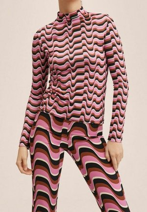 Mujer vistiendo un conjunto a juego de camiseta de manga larga y pantalones con un patrón de rayas onduladas en rosa, marrón, negro y blanco sobre un fondo neutro.