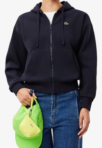 Marineblauw cropped hoodie met een rits aan de voorkant en twee zakken, voorzien van een klein groen logo. Een paar groene en gele accessoires worden in de hand gehouden.
