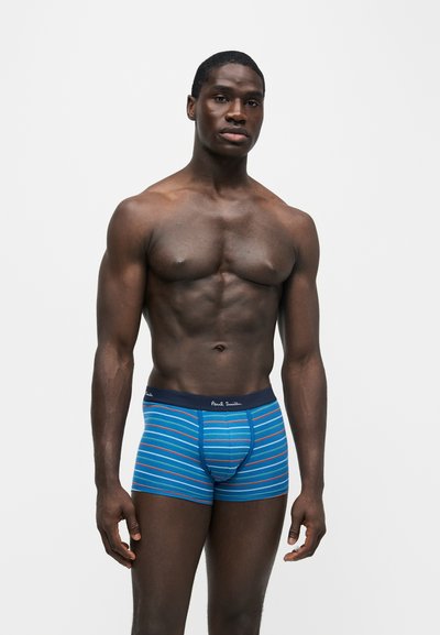Homme musclé debout devant un fond blanc portant un slip boxer bleu avec de fines rayures horizontales orange, blanche et marine.
