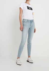 Camiseta blanca con logo negro, jeans ajustados de color azul claro y zapatillas blancas sin cordones con detalle de flecos. Tejido suave, ajuste casual.