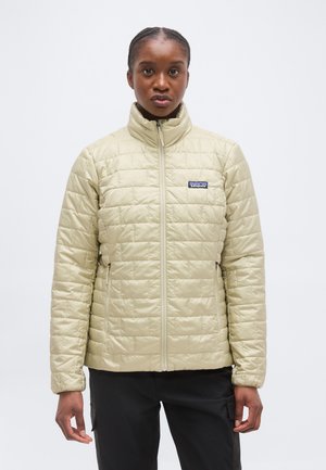 Άτομο που φοράει ανοιχτό μπεζ μπουφάν με καπιτονέ ύφασμα και ψηλό γιακά, καθώς και μαύρο παντελόνι, στέκεται μπροστά σε απλό λευκό φόντο.